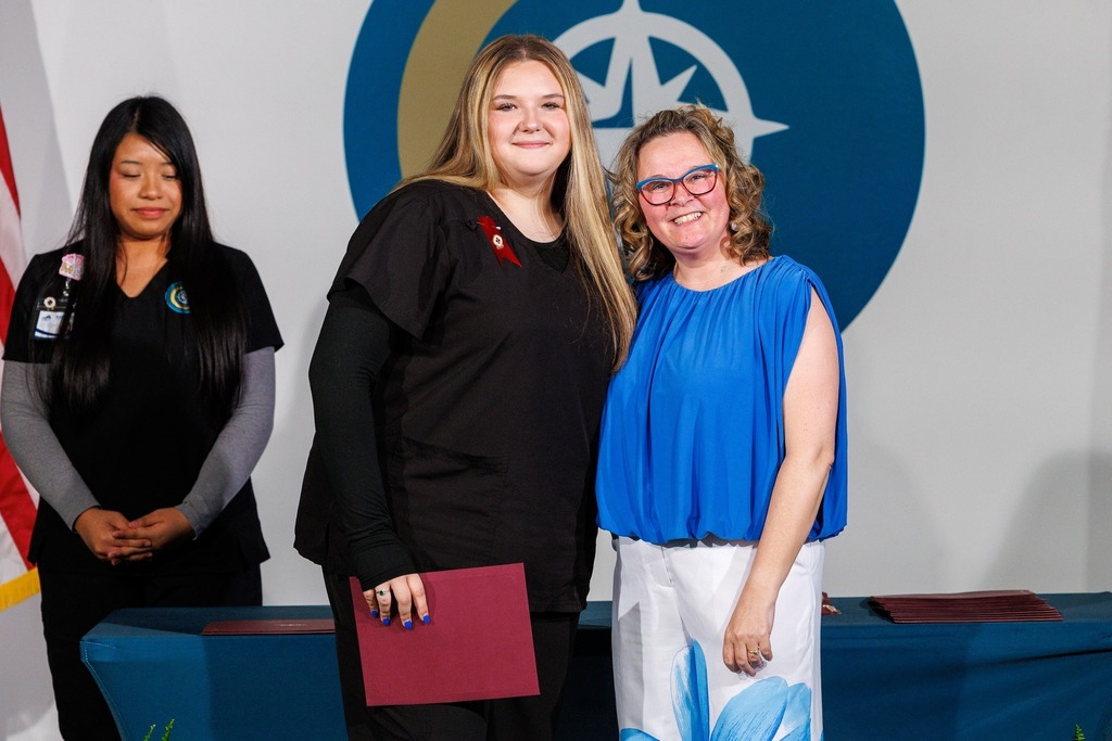Three women stand behind a blue table, one holding a folder. A woman holds a red folder, smiling. Behind them is a wall with a blue logo.