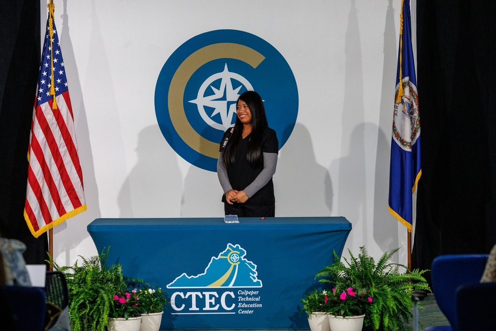 A woman stands at a podium with a blue banner reading "CTEO" behind her, next to two flags and potted plants.