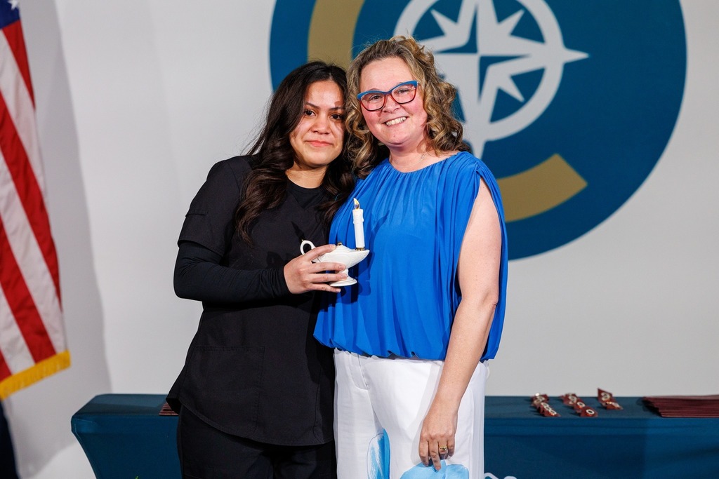 Two women stand next to each other in front of a logo and flag. One holds a candle.