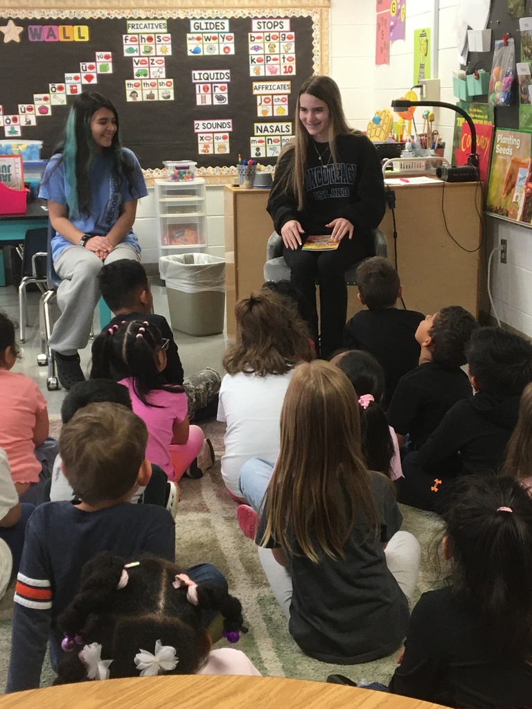Several children sit on the floor in a classroom. Two women stand in front of them, one holding a book.