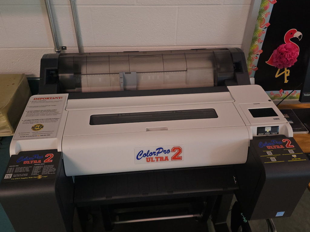 Large white printer, labeled "ColorPeel 2 Ultra 2", on a table. White wall and pink flamingo banner in the background.