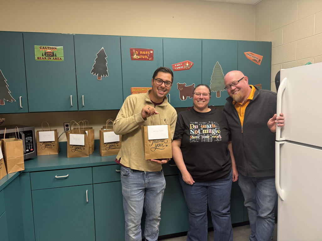Three people pose in a kitchen with teal cabinets; one holds a paper bag, others lean on a fridge.