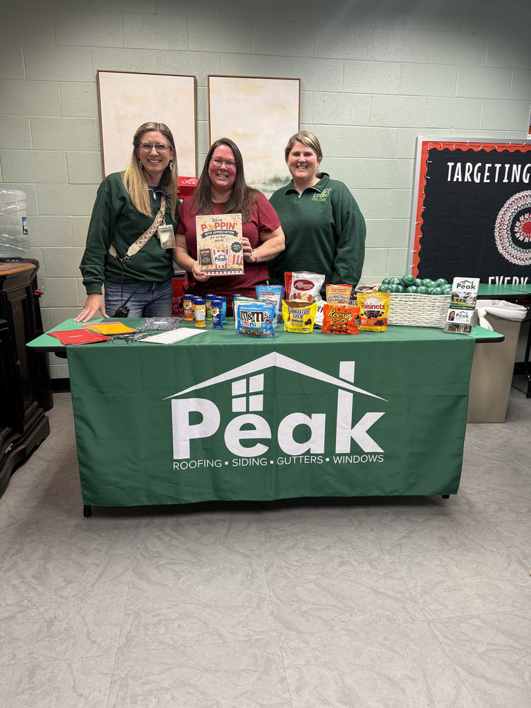 Three women in green shirts stand behind a table with Peak logo. Table displays various snacks. Background includes a classroom setting.