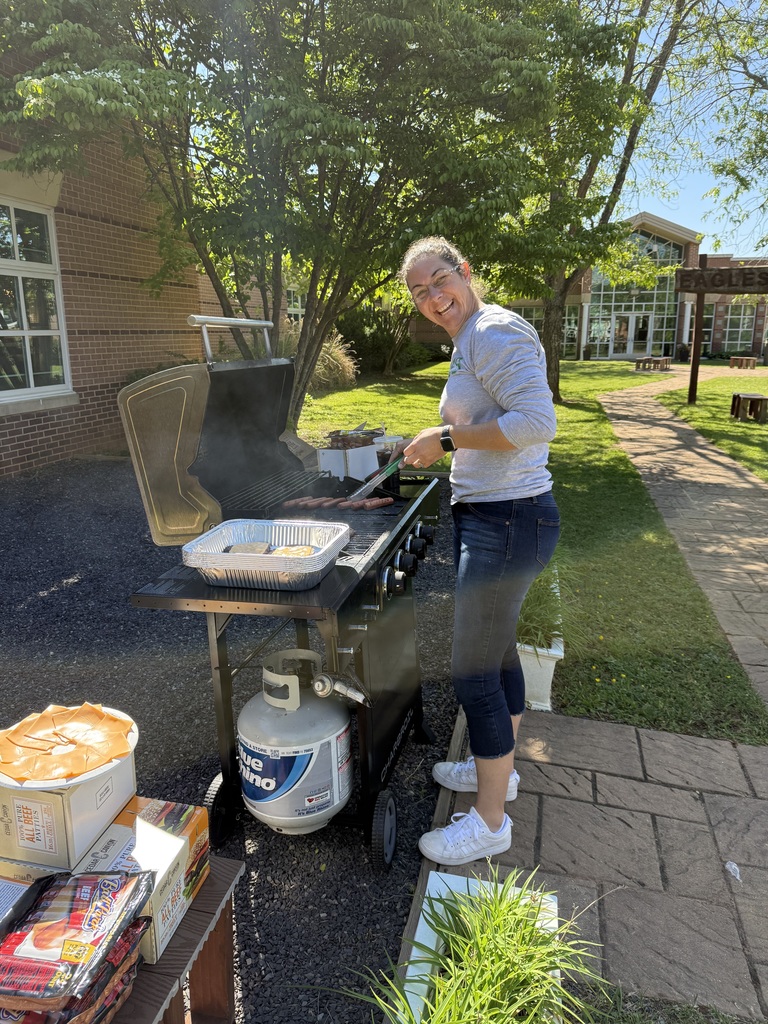A person stands outdoors, cooking food on a grill with a propane tank nearby, near a building.