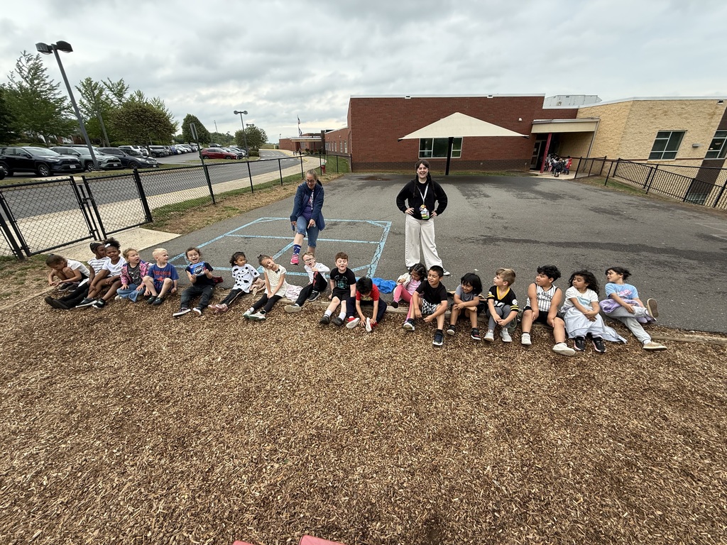 A group of children sits in a row on mulch in a school parking lot. Two adults stand behind them.