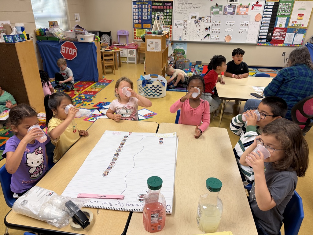 Classroom with children at desks, drinking from cups. Some hold white papers, others sit with adults.