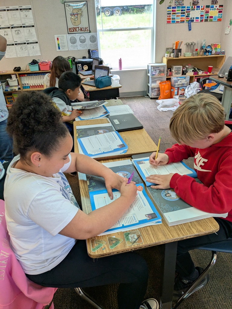 A classroom with children at desks, one girl with curly hair in white, another boy in red. Both hold pencils and write on papers.