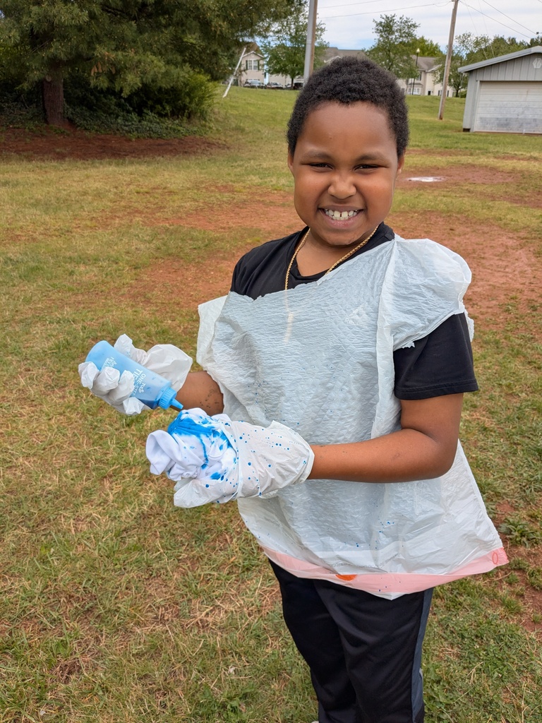 A child in a protective apron holds a bottle and a glove, standing in a grassy field.