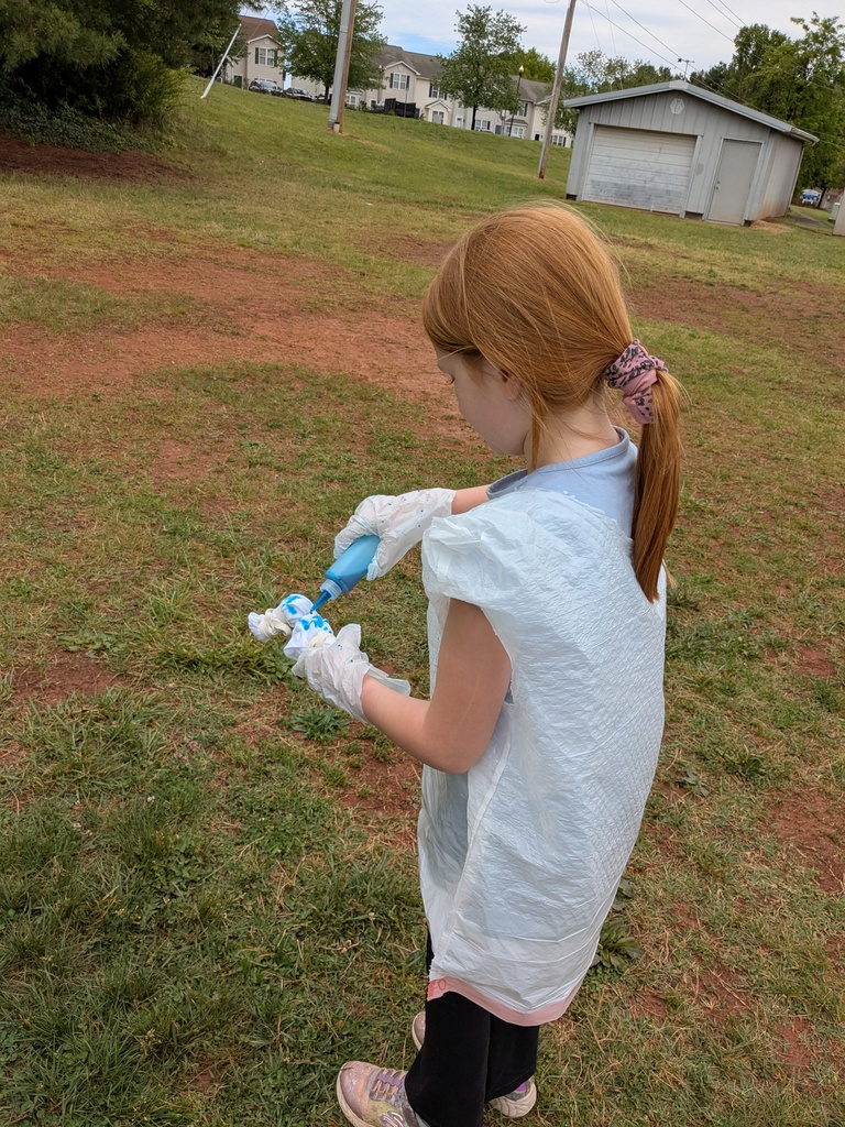 A young girl wearing gloves and an apron holds a blue object while standing on a grassy field.
