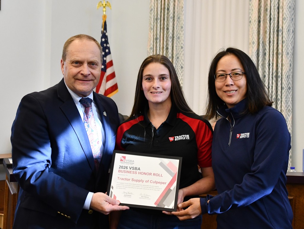 Three people stand together, presenting a certificate to a smiling woman in sportswear. A flag hangs behind them.