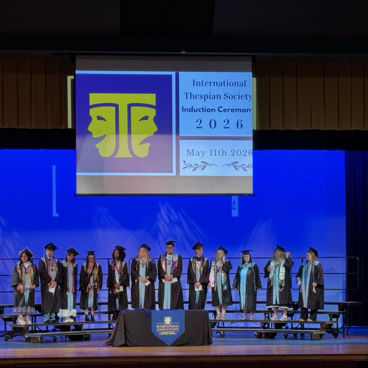 a group of students in cap and gown standing on stage risers being inducted into the International Thespian Society.