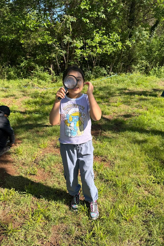Child standing on grassy hill holding magnifying glass up to face, with trees in background.