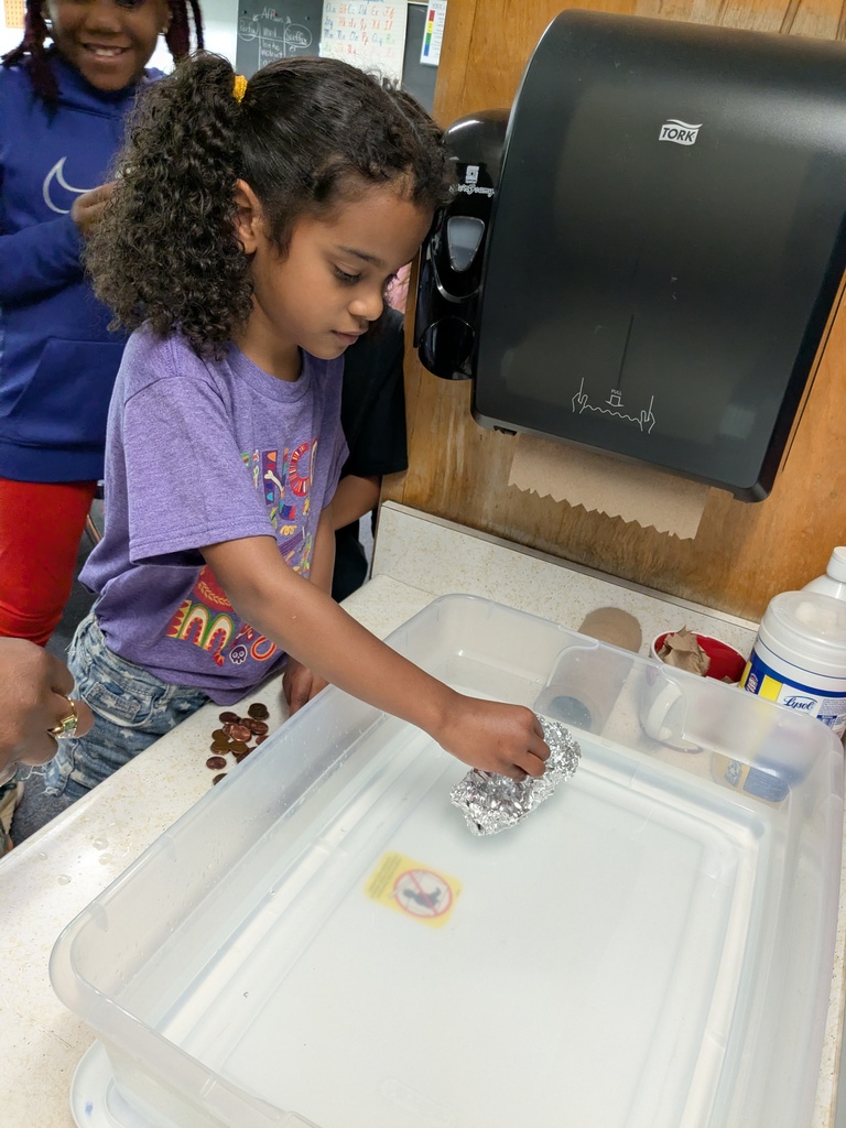 A girl with curly hair in a purple shirt examines foil in a plastic tray in a classroom.