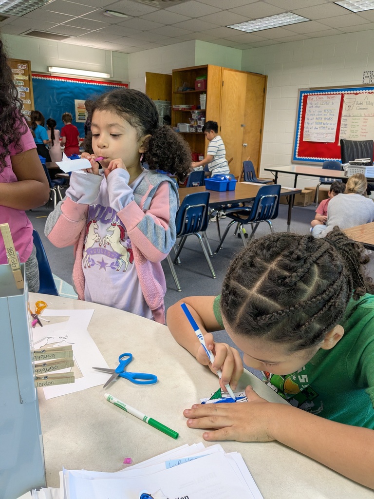 Students work on crafts in a classroom with a table full of scissors, papers, and pens.