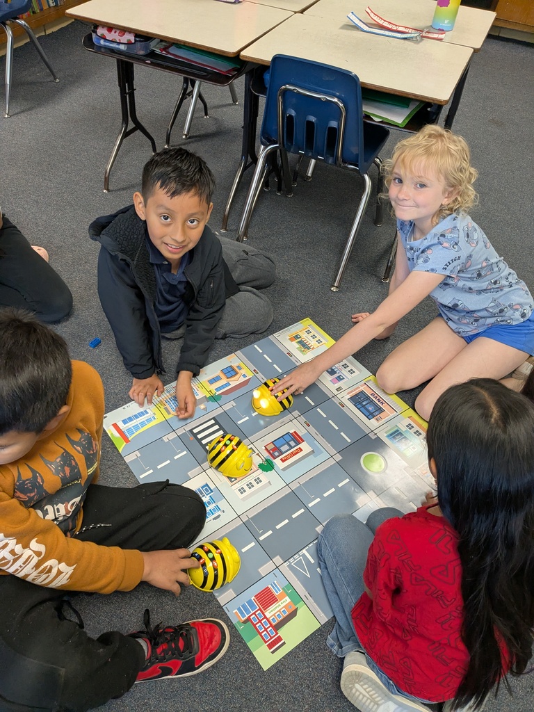 Children sit on the floor in a classroom, playing with a board game featuring roads and buildings.