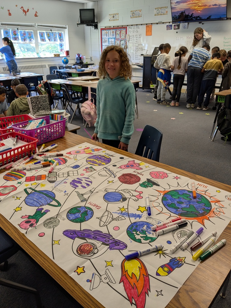 A classroom with children. A young girl stands next to a table with an art project on it.