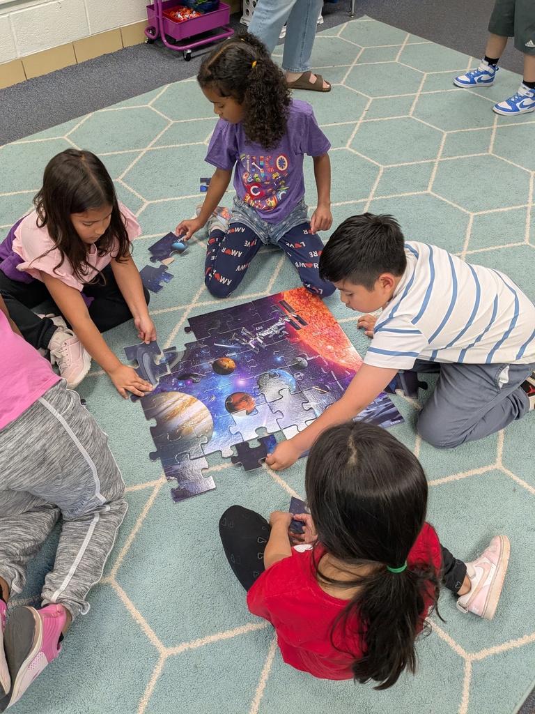 Children sit on the floor, assembling a puzzle with a space theme. A blue carpet has a hexagonal pattern.