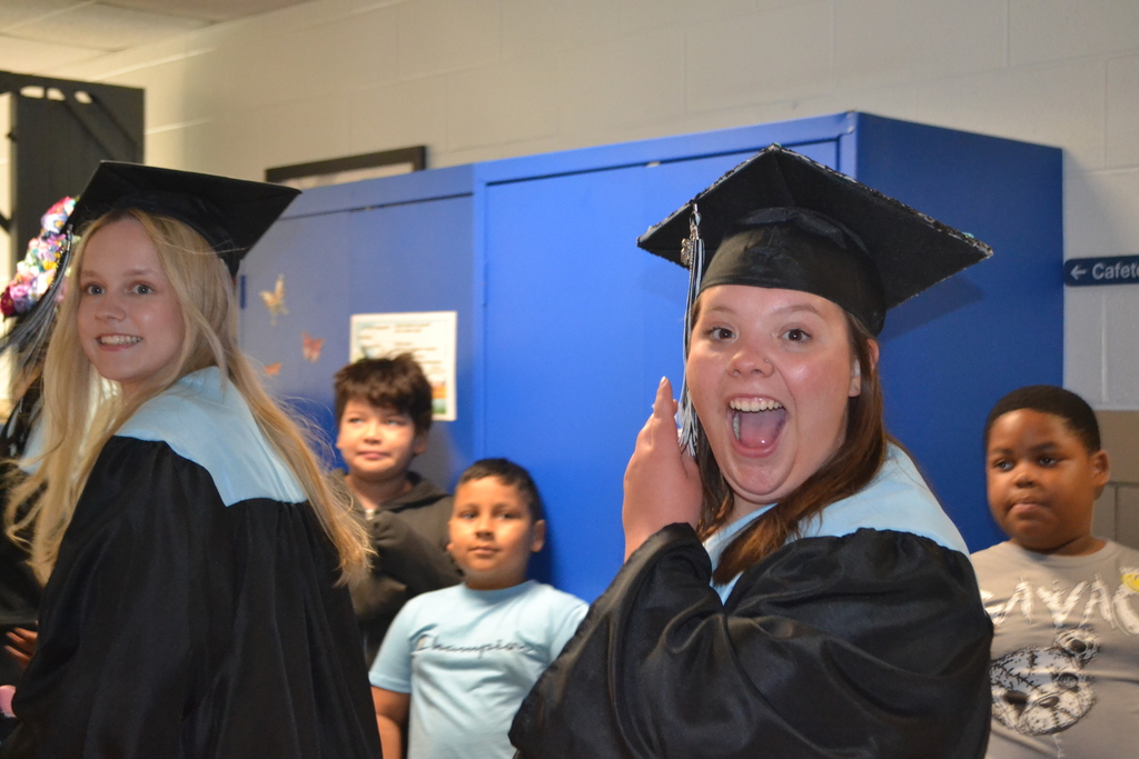Graduates in caps and gowns; one smiling and holding a cell phone. Children stand behind them. A blue locker with posters in the background.