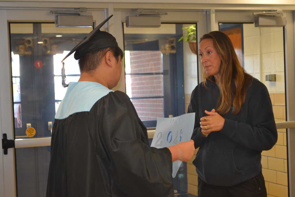 A person in a graduation gown hands a paper to a person in casual clothing in a hallway.
