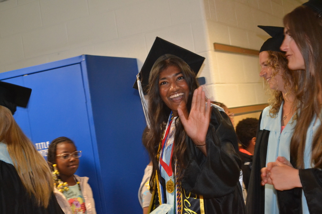 Four people in caps and gowns, smiling and talking, with one woman waving, against a white and blue wall.