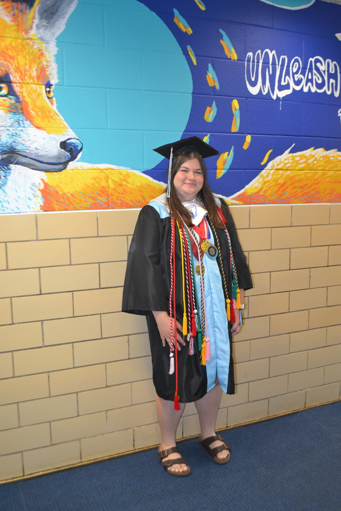 Woman in graduation gown and cap stands against a wall with a fox mural and brick pattern.