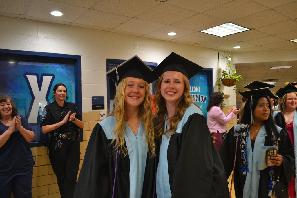 Two women in graduation robes and caps smile in a hallway. People in background wear caps and gowns, some clapping.