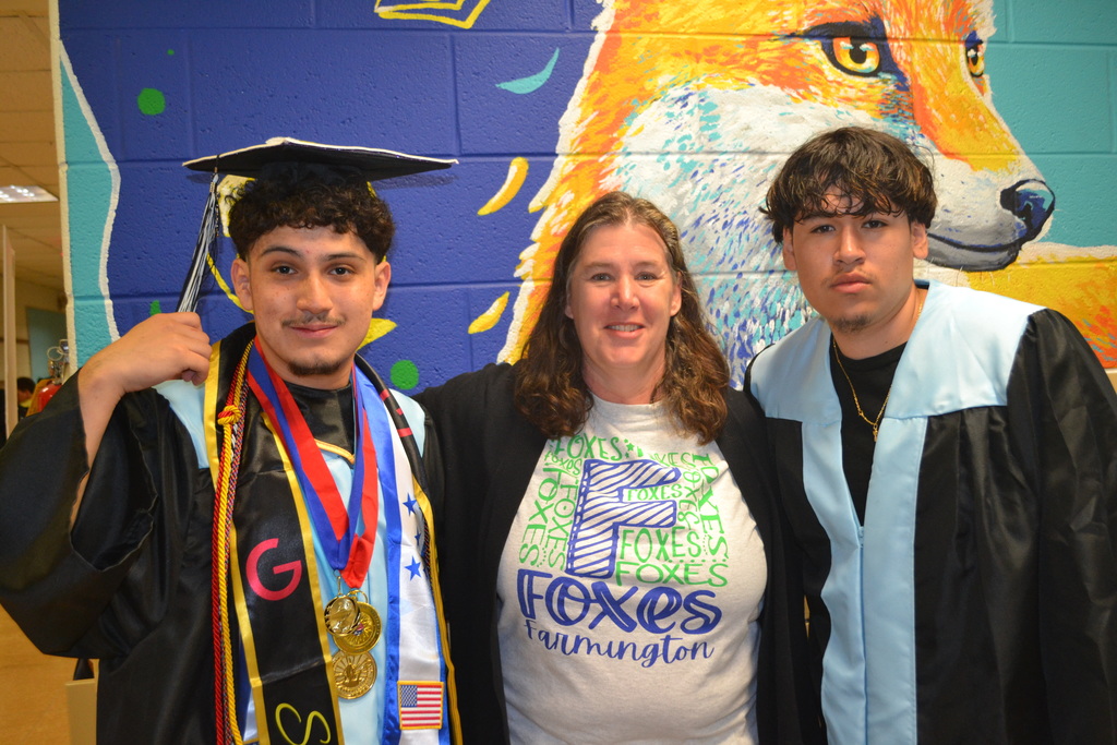 Three people in caps and gowns stand together in front of a mural of a fox.