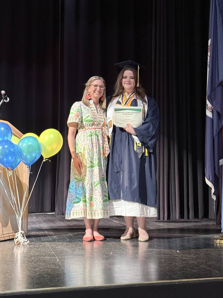 Two women on stage, one holding a diploma, in front of a dark backdrop with balloons.