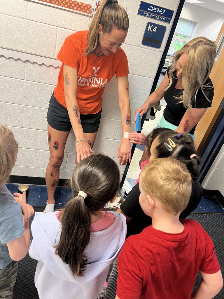 Two women help several children look at their shoes. A woman in orange and black has tattoos on her legs.