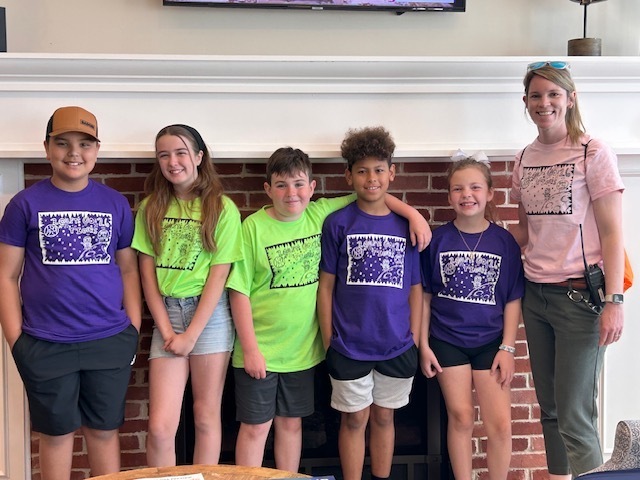 A group of six children and an adult woman, wearing matching shirts, stand in front of a brick fireplace.