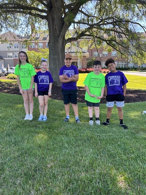 Six children stand in a grassy area, wearing matching shirts. One wears a hat. Trees and buildings are in the background.
