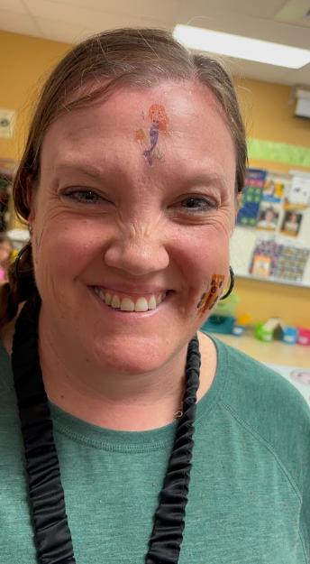 A woman with a mermaid face painting on her forehead smiles, wearing a green shirt and a black necklace.