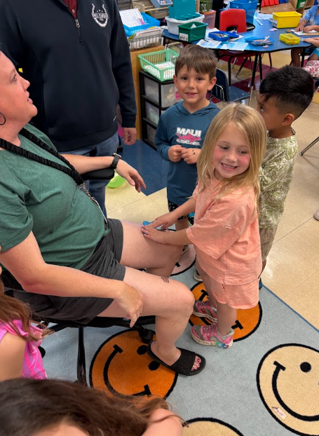 Woman sitting in chair with children; one child holding woman's hand, smiling. Background shows chairs, tables, and toys.