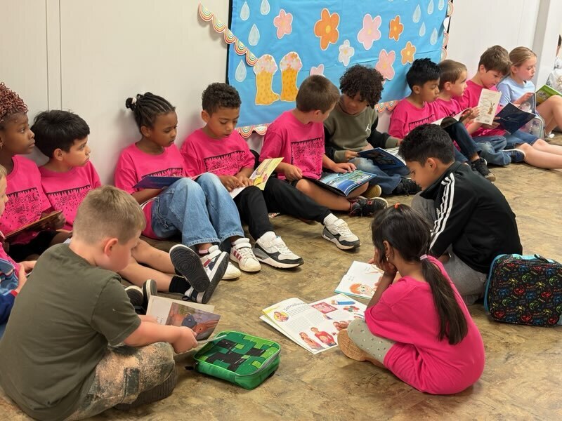 A group of children in pink shirts sit on the floor reading books; one wears a green shirt.