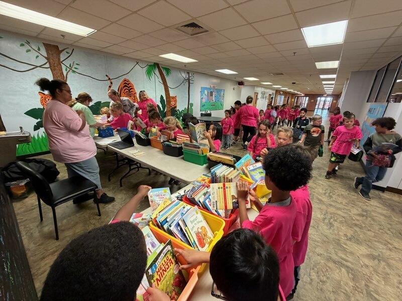 A classroom with a jungle mural, where children in pink shirts gather around tables filled with books.