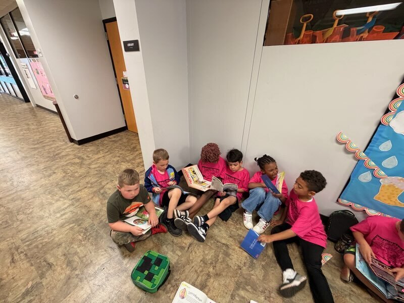Several children in pink shirts sit on the floor in a hallway, engaged with books. A colorful wall mural is behind them.