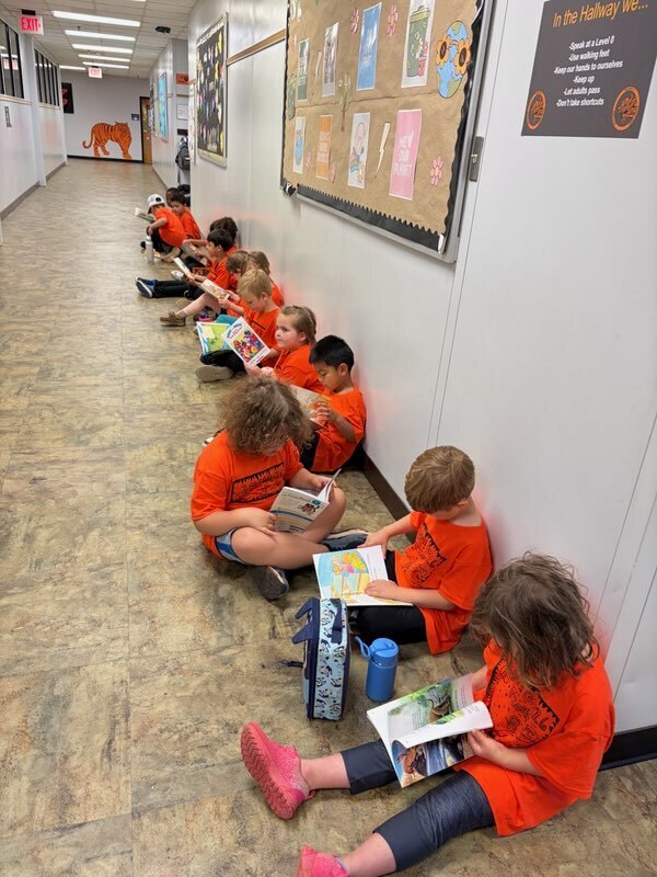 A row of children in orange shirts sit along a wall, reading books. The wall has posters.