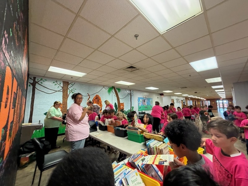 A group of children in pink shirts stand around a table with books. An adult woman stands nearby.