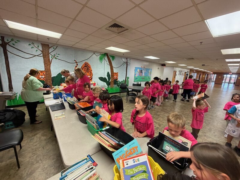 Kids in pink shirts stand around tables, some holding books, with an adult behind them. A mural is on the wall.