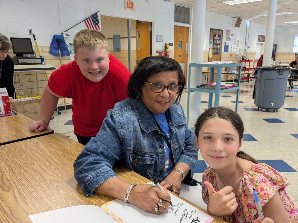 A woman with glasses and a denim jacket sits at a desk, writing. A boy in a red shirt stands behind her. A girl with a floral shirt sits next to her.