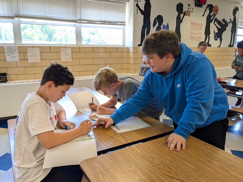 Three students in a classroom with white walls and posters. One student is drawing on paper while another looks on.