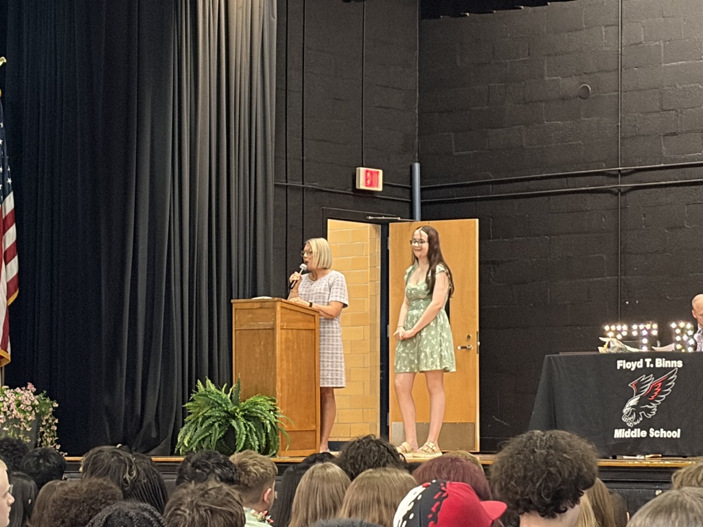     A student in a green floral dress stands beside a podium on a middle school auditorium stage while a staff member speaks into a microphone during an awards ceremony. Students and faculty are seated in the background.