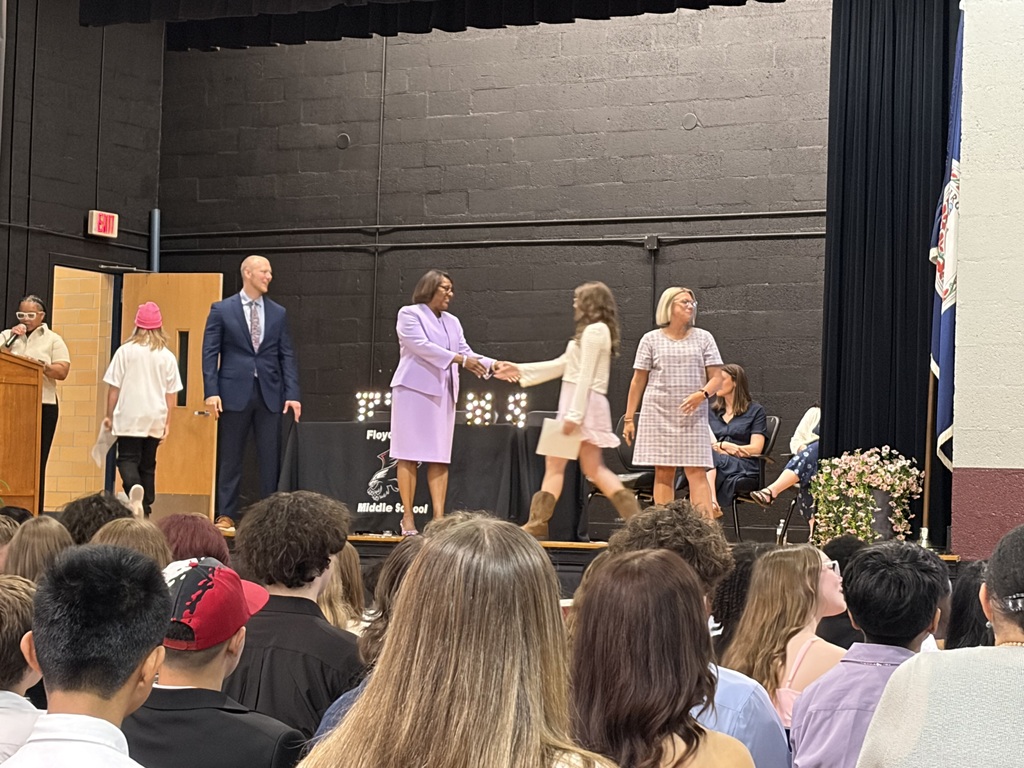     A student walks across a middle school auditorium stage to receive an award and shake hands with school administrators during a promotion ceremony. Audience members are seated in the foreground.