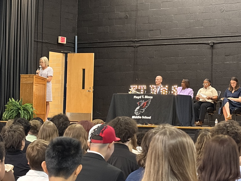     A staff member speaks at a podium during a middle school awards or promotion ceremony. School administrators are seated at a table on stage while students fill the audience.
