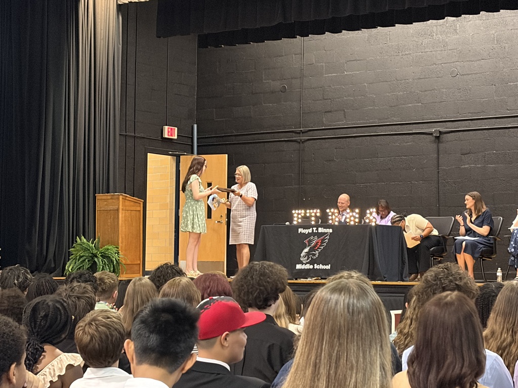     A student in a green dress receives an award or certificate from a staff member on stage during a middle school ceremony while administrators and students look on.