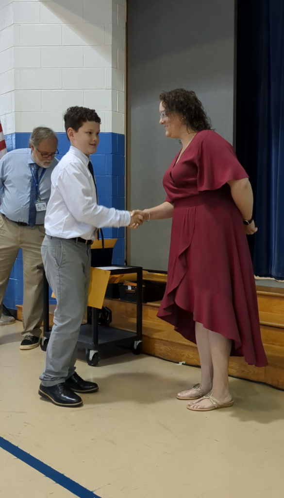 A person in a white shirt and pants shakes hands with a person in a maroon dress. Behind them, another individual in a blue shirt and tie watches.