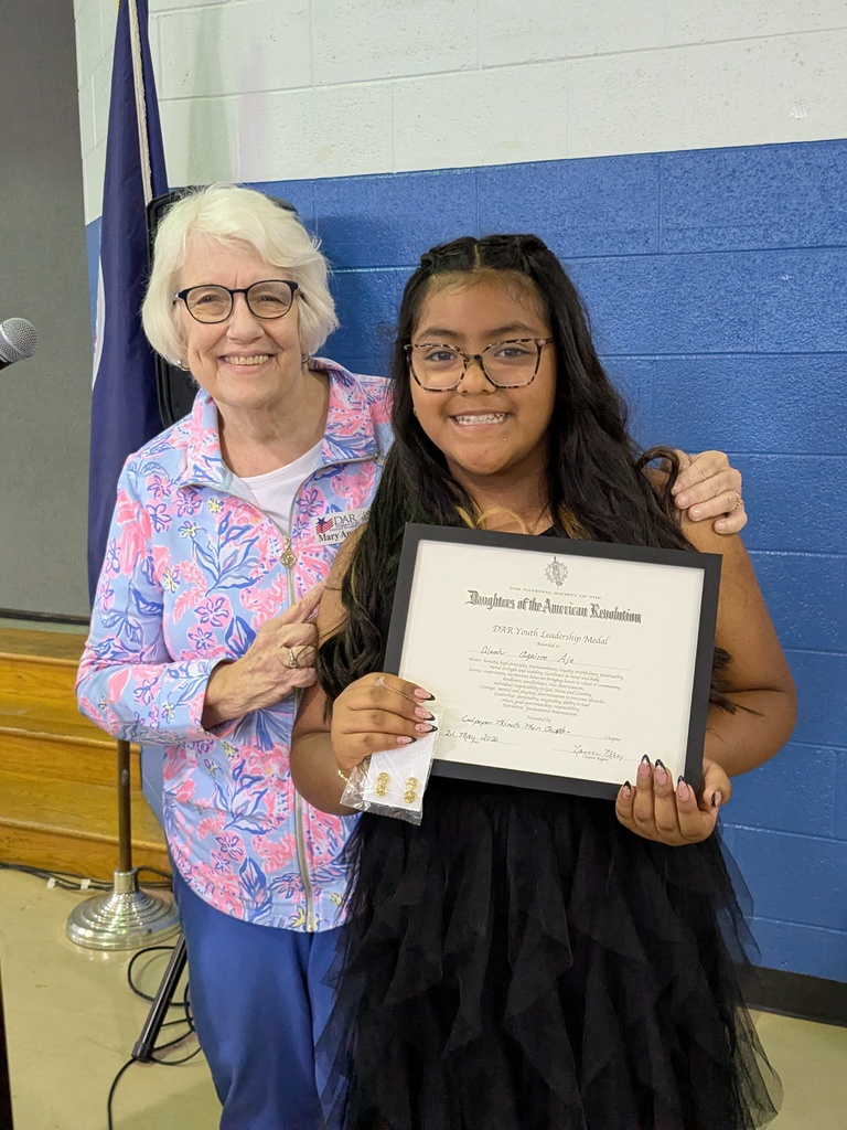 A woman in glasses holds a certificate, standing with a girl in a black dress. A flag and microphone are in the background.