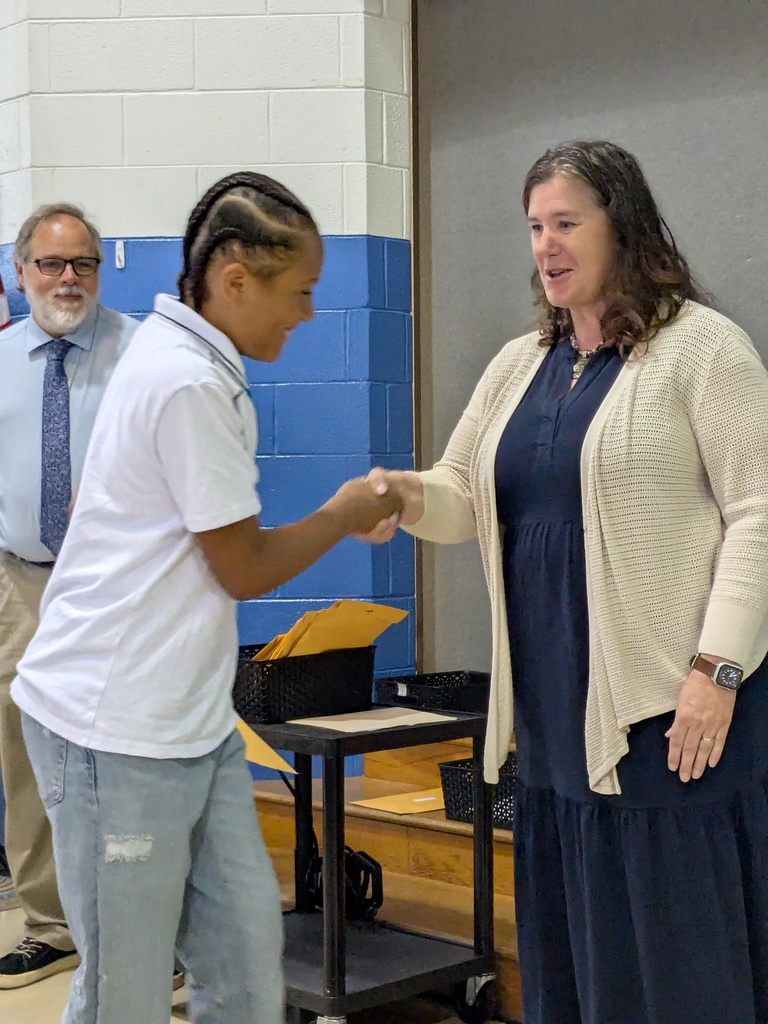 A student and a teacher shake hands in a school hallway. Behind them, a man in glasses and a tie stands.