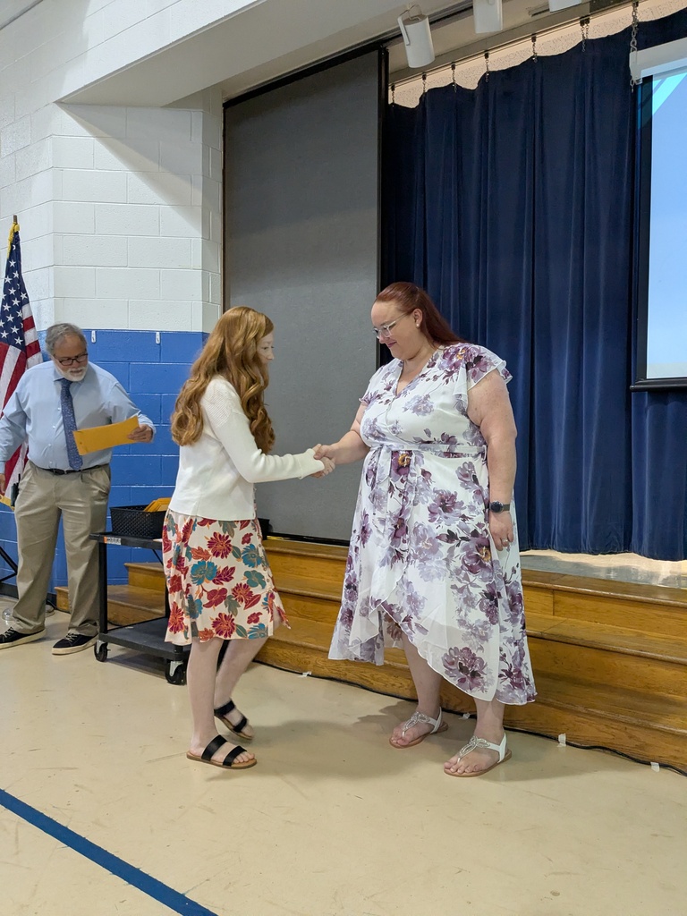 Two women shake hands in a classroom. One wears a white top and floral skirt. The other wears a floral dress. A man stands behind them holding a folder.