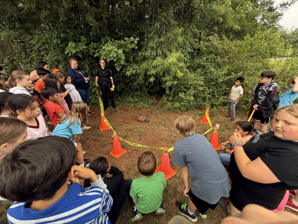 A group of elementary students gathered outside around a turtle laying eggs in a small roped-off area near the playground. Orange safety cones and caution tape surround the turtle while students and staff observe quietly from a safe distance. Trees and greenery fill the background as the students excitedly watch the special nature moment.